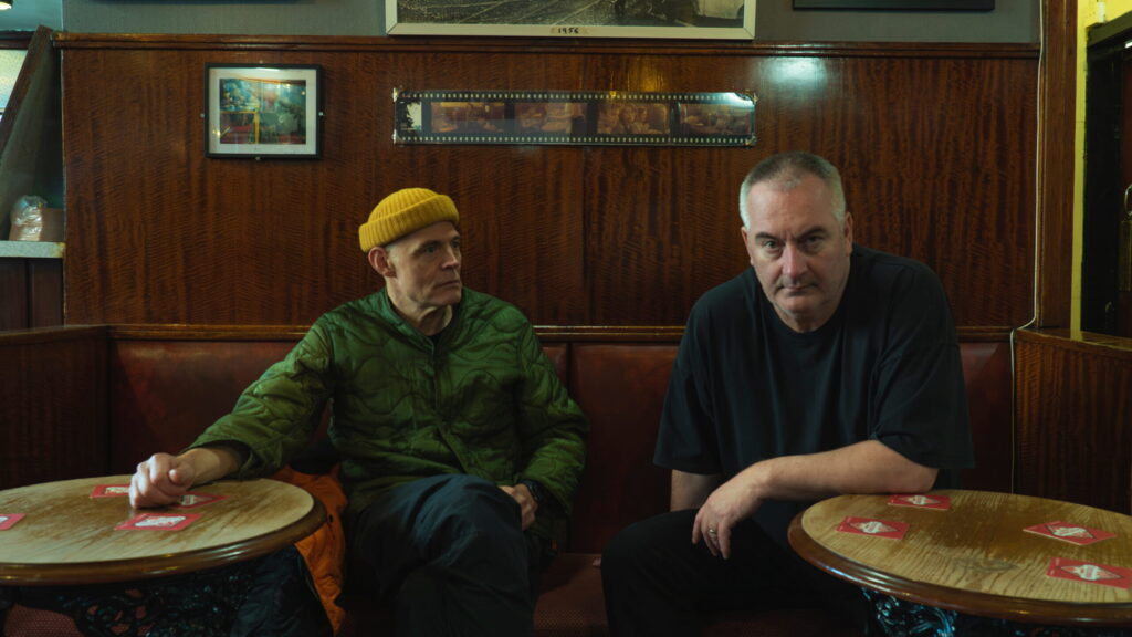 Portrait du duo électronique Slam, composé de Stuart McMillan et Orde Meikle, assis à une table dans un décor de bar boisé.