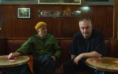 Portrait du duo électronique Slam, composé de Stuart McMillan et Orde Meikle, assis à une table dans un décor de bar boisé.