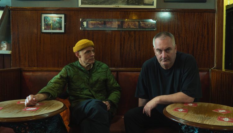 Portrait du duo électronique Slam, composé de Stuart McMillan et Orde Meikle, assis à une table dans un décor de bar boisé.